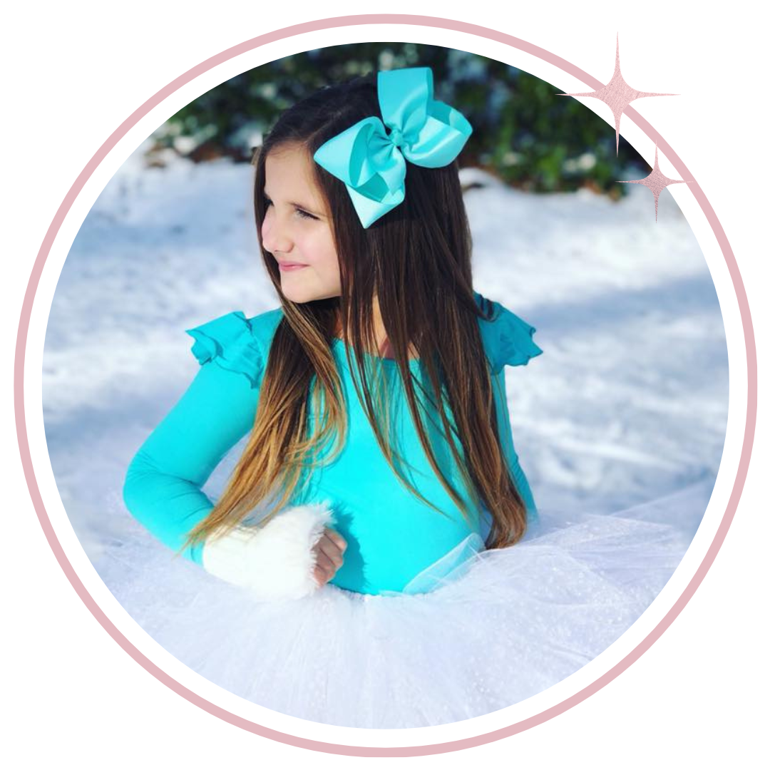 A young girl with long brown hair wearing a bright turquoise long-sleeve leotard and a matching bow in her hair, sitting in the snow. The photo is framed inside a pink circular border with sparkles in the top right corner.