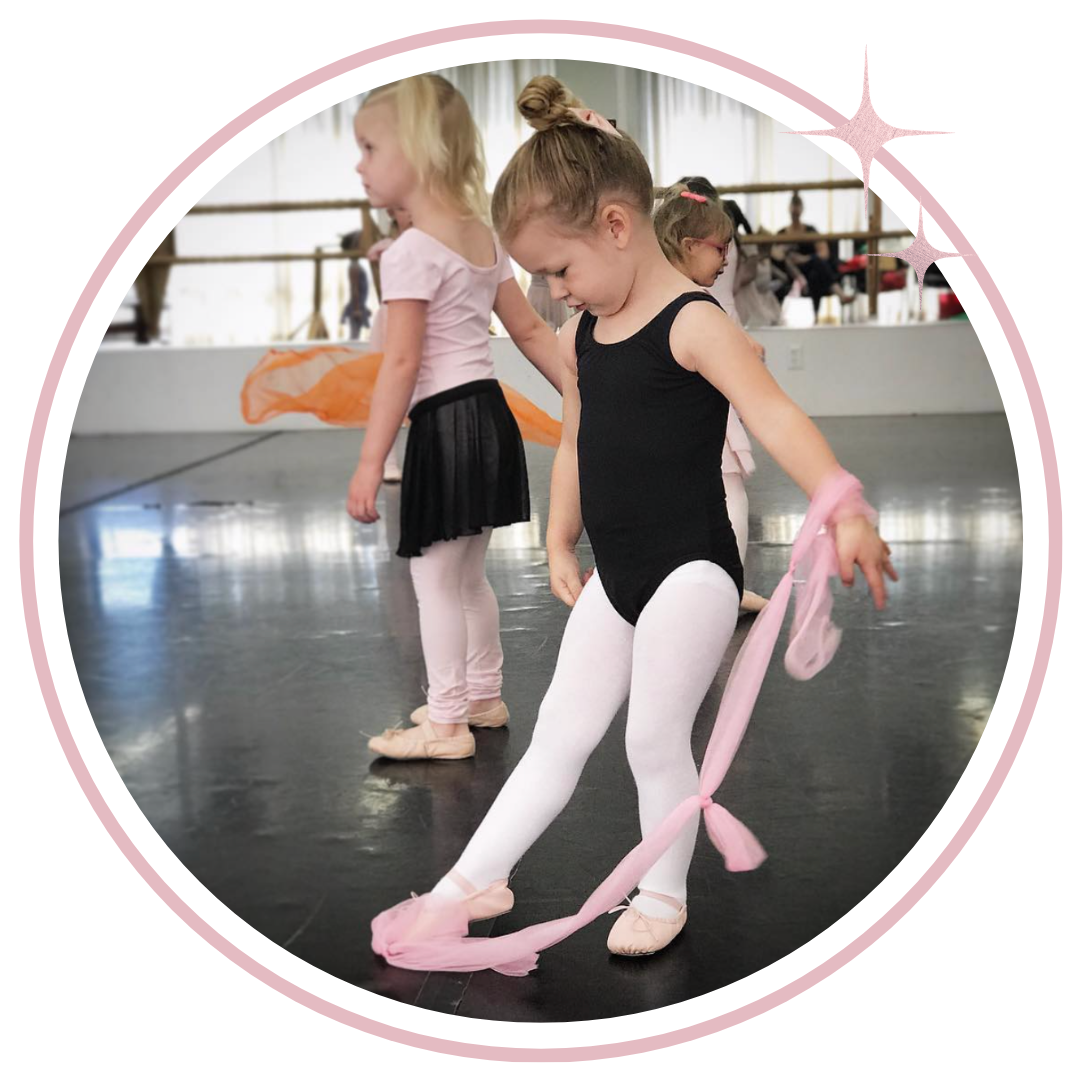 A young girl in a black leotard and white tights practices ballet with a pink ribbon in a dance studio, framed within a pink circular border.